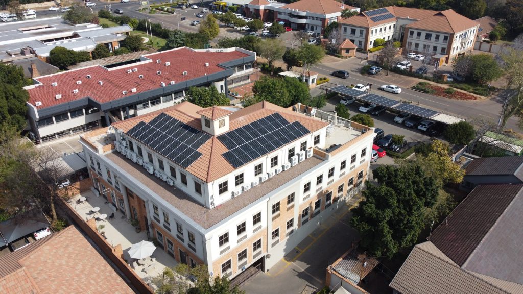 Aerial view of a commercial office building with rooftop solar panel installation in an urban area, showing multiple solar arrays and surrounding parking lot.